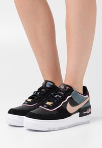 Svarta mocka- och lädersneakers med rosa detaljer, vit mellansula och grå hälpatch. Har guldbranding och perforerade tår.