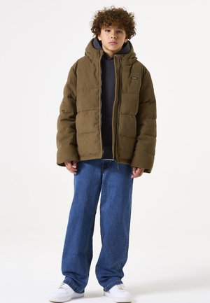 Chaqueta de puffer de pana marrón con capucha, con cierre de cremallera en el frente, llevada sobre una sudadera gris oscuro y combinada con unos jeans sueltos azules y zapatillas blancas.