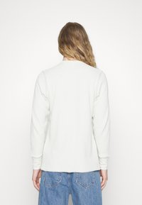 Vit långärmad skjorta med texturerad, ribbad design; rund halsringning; något böjd fåll baktill; kombinerad med blå högmidjade jeans.