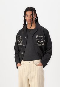 Black jacket with white animal graphics on front, button closure, and curved hem, paired with a dark sweater and cream-striped pants.