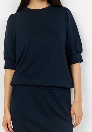 Vrouw draagt een marineblauwe jurk met korte pofmouwen en een aansluitende rok, poseert tegen een effen witte achtergrond.