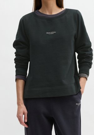 Personne portant un sweat-shirt noir Marc O’Polo et un pantalon sombre, debout avec une main dans la poche et l'autre le long du corps.