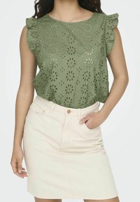 Top verde oliva con motivo floreale a occhio, maniche a volant; abbinato a una gonna di jeans beige chiaro con bottoni frontali e tasche.