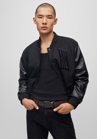 Veste bomber noire avec des manches en cuir et un logo brodé. Portée sur un débardeur noir, associée à un pantalon texturé noir et une ceinture marron.