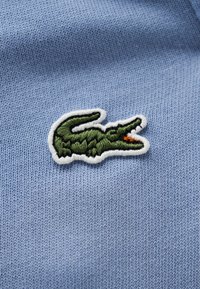 Πράσινο κεντημένο λογότυπο κροκοδείλου με ανοιχτό στόμα, ραμμένο σε ανοιχτό γαλάζιο πλεκτό ύφασμα.