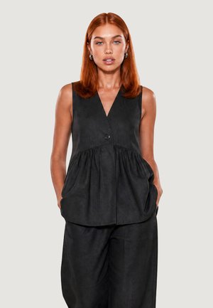 Donna con capelli rossi che indossa un top nero senza maniche con bottoni e pantaloni larghi abbinati, in piedi con le mani nelle tasche su uno sfondo uniforme.