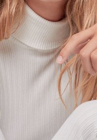 Kobieta w białym swetrze z ryżowym golfem, z jasnobrązowymi włosami, delikatnie dotykająca swoich pasm.