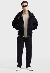 Geacă neagră din blană artificială cu fermoar și accente din piele, purtată peste un tricou bej și asortată cu pantaloni închiși la culoare și adidași albi.
