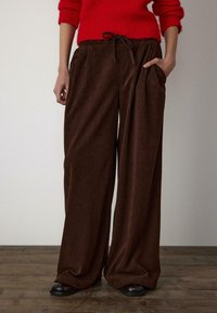 Brązowe szerokie spodnie ze sztruksu z pionową fakturą pr�ążkowaną, elastycznym pasem i bocznymi kieszeniami, zestawione z czerwonym dzianinowym swetrem.