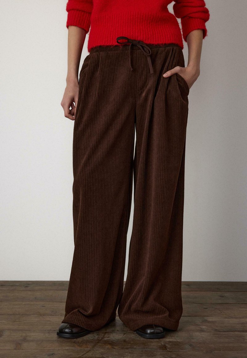 Brązowe szerokie spodnie ze sztruksu z pionową fakturą prążkowaną, elastycznym pasem i bocznymi kieszeniami, zestawione z czerwonym dzianinowym swetrem.