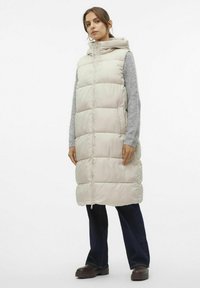 Gilet long léger rembourré en beige clair avec une capuche, comportant une fermeture éclair complète, des poches latérales et des manches de pull texturé gris.