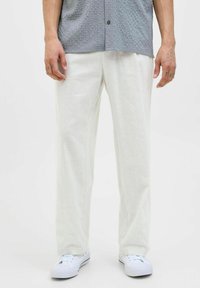 Pantaloni de culoare deschisă, cu croială lejeră, dintr-un material texturat, asortați cu adidași albi și o cămașă gri cu model cu mânecă scurtă.