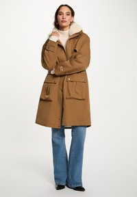 Parka marrone con cappuccio foderato in pelliccia sintetica, zip frontale, due ampie tasche a pattina e texture morbida. Indossata sopra un maglione crema e dei jeans.