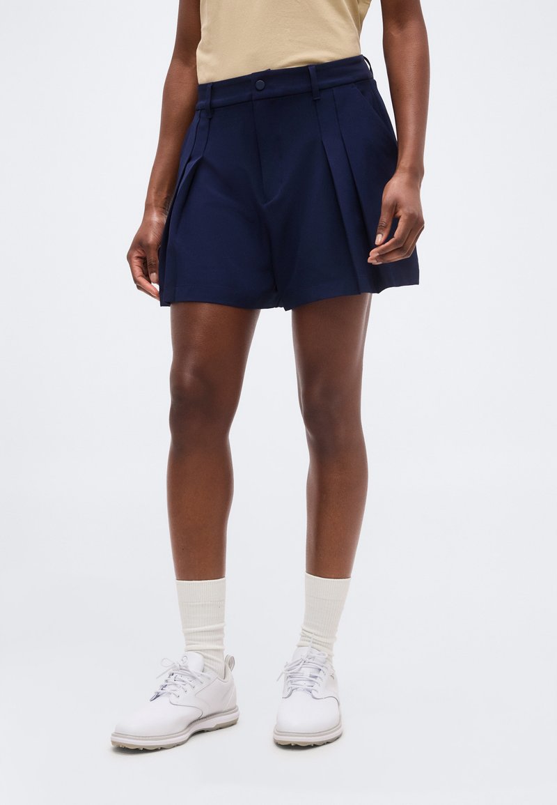 Personne portant un short bleu marine sur mesure avec des plis, un haut beige, des chaussettes blanches montantes et des chaussures blanches à lacets, debout devant un fond uni.