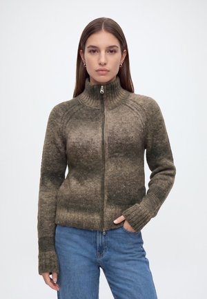 Jeune femme portant un pull en maille zippé dégradé marron et vert et un jean bleu, debout avec une main dans la poche devant un fond uni.