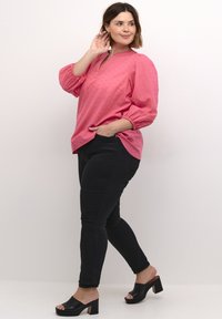 Blusa rosa con un motivo strutturato, maniche arricciate e scollo a V. Abbinata a pantaloni neri aderenti e tacchi aperti neri.