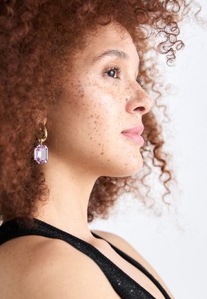 Gouden oorring met een transparante paarse edelsteen. Het model heeft krullend rood haar en sproetjes, en draagt een zwarte top met een gestructureerd oppervlak.