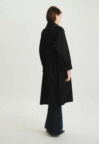 Manteau noir en trench avec une taille ceinturée, des manches larges et un col ; texture lisse ; porté sur un pantalon en denim évasé.