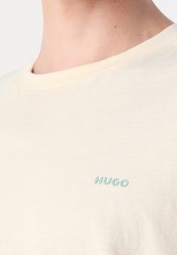 T-shirt en coton beige avec un col rond et un logo "HUGO" bleu clair sur la poitrine, présentant une texture lisse et un design simple.