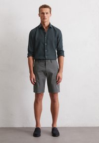 Shorts gris avec une coupe décontractée associés à une chemise à boutons vert foncé. La chemise a des manches retroussées et une texture douce. Chaussures noires.