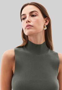 Top sin mangas de color verde oscuro con cuello alto en tejido de punto acanalado. La textura es suave, con un cuello alto y costuras mínimas. Pendientes de oro y plata.