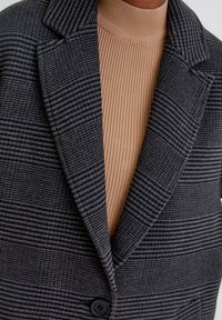 Blazer grigio a scacchi con rever a punta e un unico bottone nero, sovrapposto a un maglione beige a collo alto a coste.