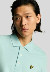 Lichtblauw katoenen poloshirt met een geel geborduurd logo op de linkerborst, voorzien van een kraag en een knoopsluiting met drie knopen.