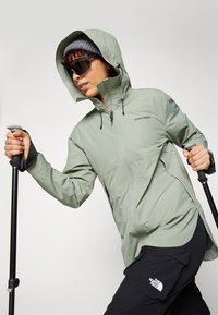Giacca impermeabile verde chiaro con cappuccio, zip frontale e lacci regolabili. Il modello tiene in mano dei bastoni da trekking e indossa pantaloni scuri e occhiali da sole.