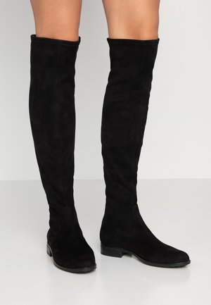 Bottes en daim noir montant jusqu'aux genoux, portées sur des jambes nues, avec des talons blocs bas et des bouts arrondis, posées sur un sol gris clair.