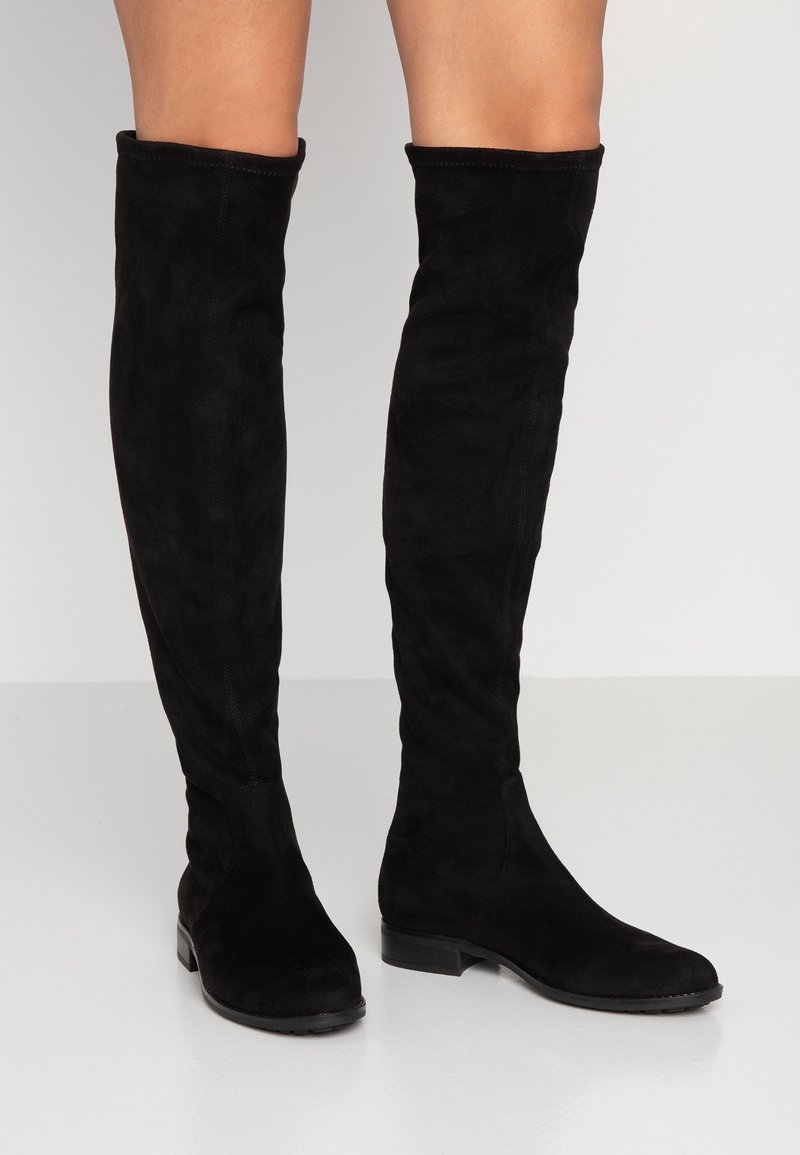 Bottes en daim noir montant jusqu'aux genoux, portées sur des jambes nues, avec des talons blocs bas et des bouts arrondis, posées sur un sol gris clair.