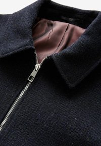 Jachetă din lână bleumarin cu o închidere cu fermoar, guler ascuțit și captuseală interioară netedă. Exterior texturat cu un model de țesătură subtil.
