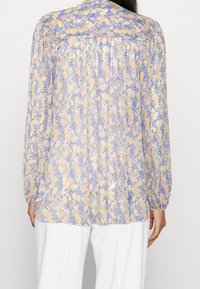Blouse légère à manches longues avec un motif floral pastel aux nuances de violet, jaune et rose. Comprend des plis et un ourlet arrondi.
