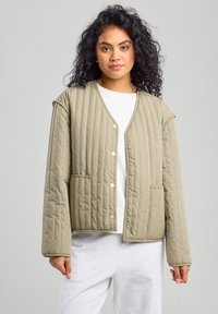 Chaqueta acolchada de color verde oliva con cuello en V, cierre frontal de botones a presión, dos bolsillos laterales y ajuste relajado. Tejido suave y texturizado con costuras en contraste.