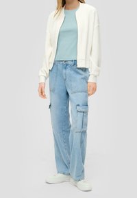 Giacca bomber bianca con texture, camicia azzurro chiaro e jeans cargo azzurro chiaro con tasche a toppa. Sneakers bianche completano l'outfit.