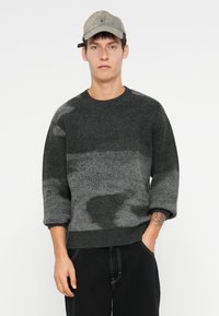 Γκρι πουλόβερ πλεξίματος με υφασμάτινο σχέδιο και τονικά μοτίβα. Το μοντέλο φοράει ένα ανοιχτό γκρι καπέλο και μαύρο παντελόνι, με τα χέρια στις τσέπες.