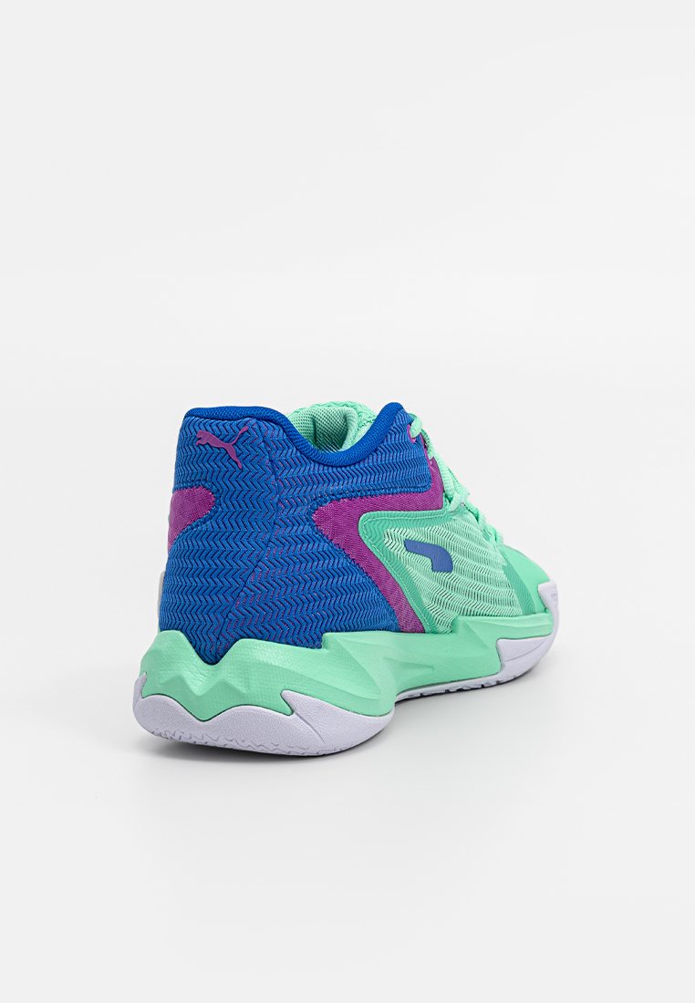 Zapatilla deportiva con parte superior de malla verde, acentos azules, patrones texturizados y suela de un ligero color púrpura. Detalle notable del logo en rosa.