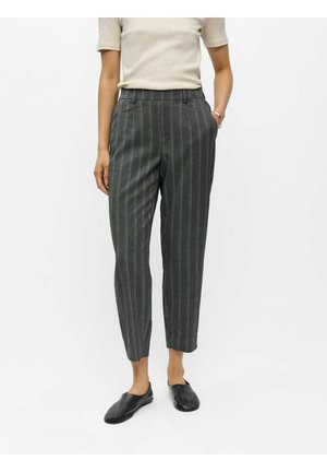 Personne portant un pantalon gris à rayures longueur cheville, un haut beige à manches courtes et des chaussures noires sans lacets, debout avec une main dans la poche.