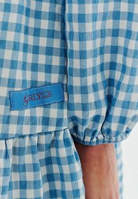 Modrá a biela károvaná látka s naťahovacími elastickými manžetami. Obsahuje malú modrú nálepku s nápisom "GRLYtude" v červenej písme.