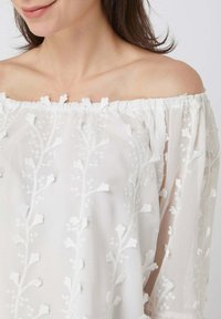 Haut blanc à épaules dénudées avec broderie florale, présentant des motifs texturés et un col élastique froncé. Matière transparente avec un design fluide.