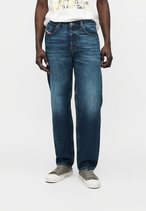 Bărbat purtând blugi largi albaștri și adidași gri, stând în picioare pe un fundal simplu și deschis, cu mâinile relaxate pe lângă corp.