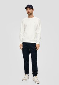 Witte sweatshirt, marineblauwe cargobroek en witte sneakers met beige accenten. Het model draagt een marineblauwe pet en staat tegen een grijze achtergrond.