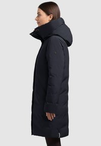 Abrigo de plumas azul marino con collar alto y capucha, que cuenta con bolsillos laterales y una tela suave y texturizada. Líneas de diseño simples y modernas.