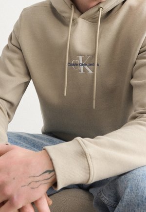 Osoba ubrana w beżową bluzę z kapturem Calvin Klein Jeans z sznurkami oraz jasnoniebieskie dżinsy, spoczywająca dłonie z małym tatuażem na lewym nadgarstku.