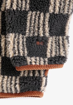 Kind met krullend rood haar draagt een rode gebreide pet en een zwart-beige geblokt fleecejack met knopen en bruine corduroy-bies.