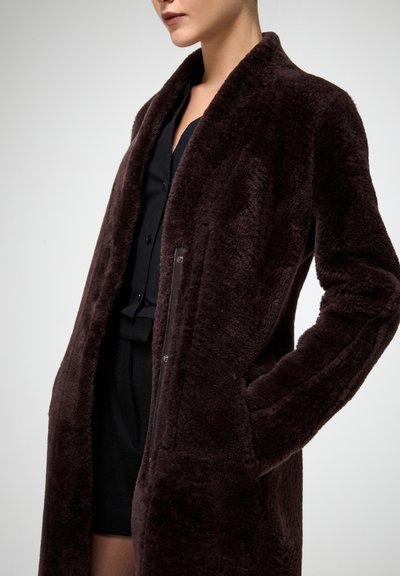 Garš, tumši brūns mākslīgās kažokādas mētelis ar šāla apkakli, sānu kabatām un mīkstu, teksturētu virsmu. Stilizēts pār melnu topu.