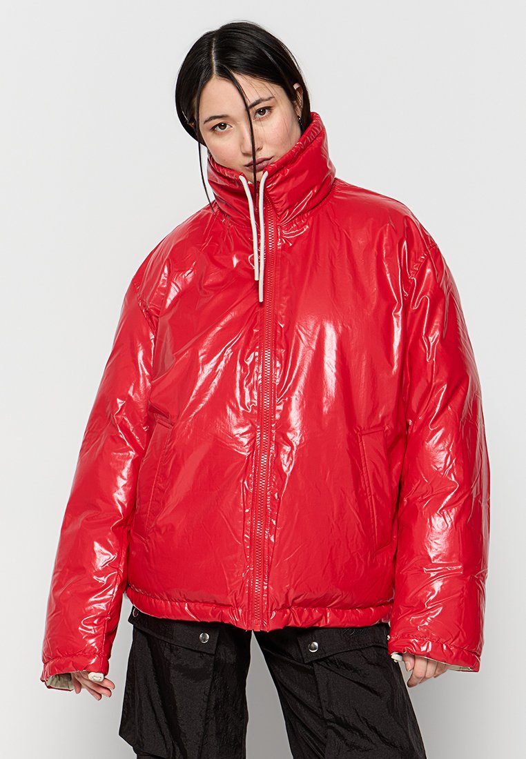 Chaqueta acolchada roja brillante con un alto cuello, cremallera frontal y detalle con cordón. Presenta un corte holgado y una textura suave y brillante.