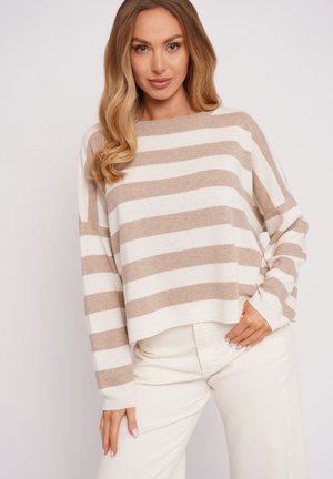 Kobieta o długich blond włosach, ubrana w beżowo-biały sweter w poziome paski i białe spodnie, pozująca na jednolitym tle.