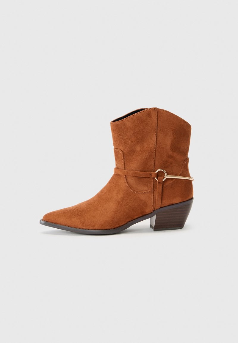 Bottine en daim marron avec petit talon empilé et bride décorative avec anneau métallique doré sur le côté, sur fond uni gris clair.