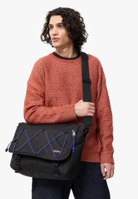 Musta viestintälaukku, jossa on säädettävä olkahihna, siniset naruakkentit ja vetskarella suljettava tasku, kiinnitetty teksturoituun kangaspintaan.