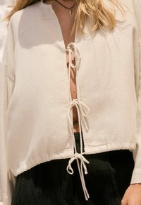 Licht beige cropped top van een gestructureerde stof, met een knooploze voorkant met strikjes, lange mouwen en een relaxte pasvorm.